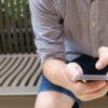 man wearing brown and white plaid sport shirt sittings on brown bench and using smartphone during day